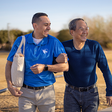 A Visiting Angels caregiver lends an arm for support to a senior male as they walk in the park.